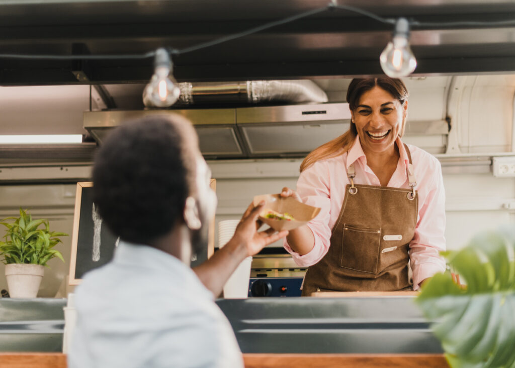 A smiling food truck owner hands a customer lunch.