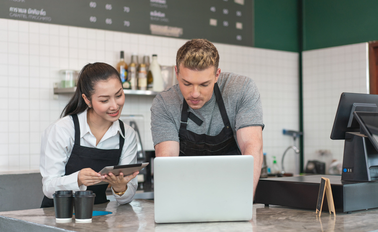 Two baristas in a coffee shop looking at a laptop and tablet, collaborating on tasks behind the counter.