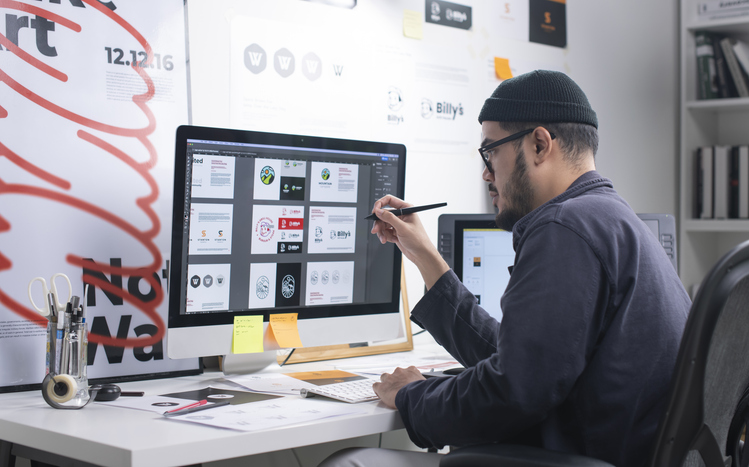 A designer in a black beanie works on a desktop computer, reviewing logo designs in a creative, modern office space.