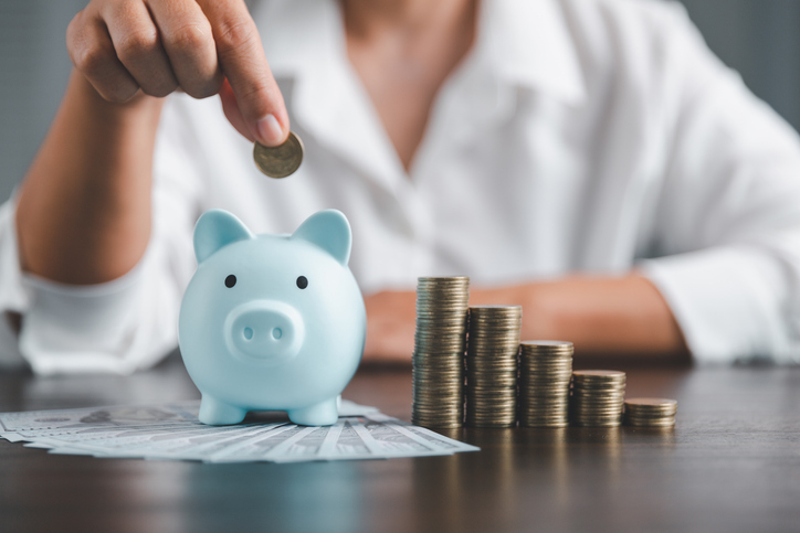 Person saving money by placing a coin into a blue piggy bank, with stacked coins and cash on a table in the foreground.