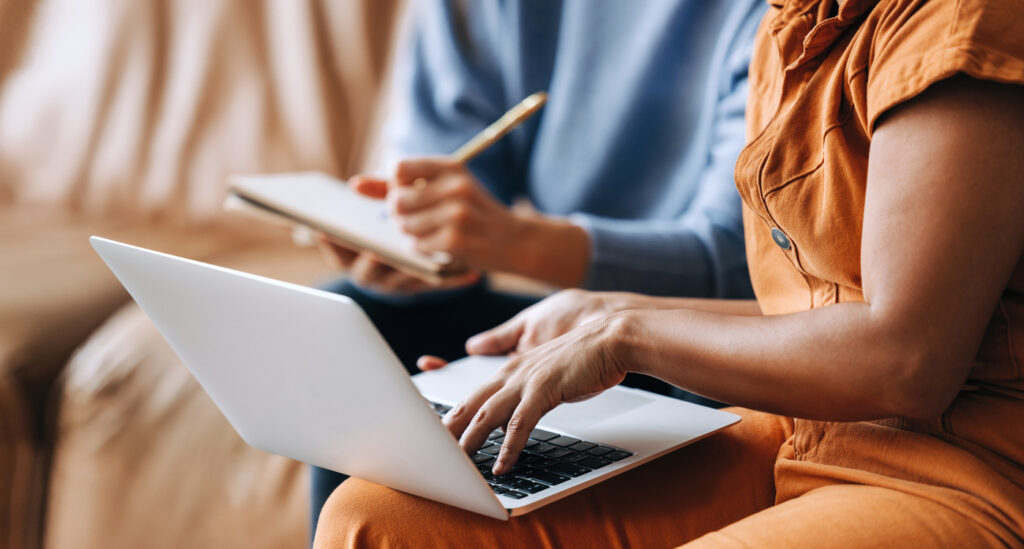 Two business women collaborate over a laptop and notebook.