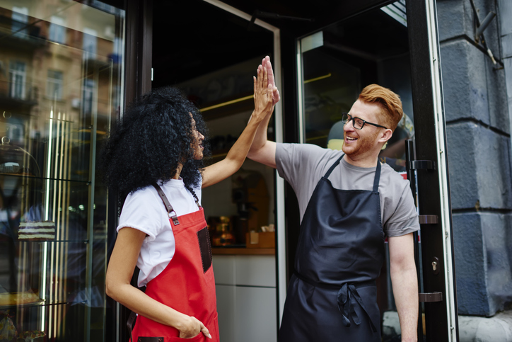 Two happy café employees in aprons high-five outside their shop, celebrating success and teamwork with smiles.