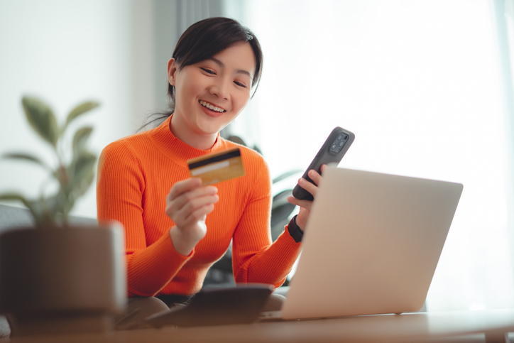 A smiling woman in an orange sweater uses a laptop, holding a credit card and smartphone, managing online banking at home.