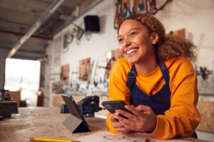 A smiling young business owner holds a tablet with a workshop behind her.