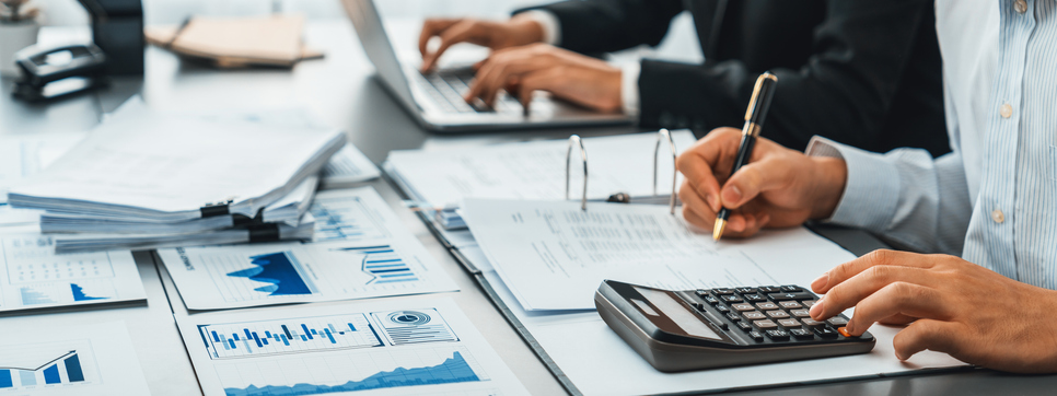 Business professionals working at a desk with financial documents, a calculator, and laptops, analyzing charts and data.