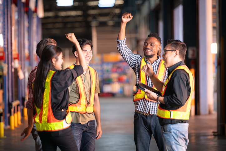 A group of warehouse employees in safety vests celebrating success together with raised hands and smiles.