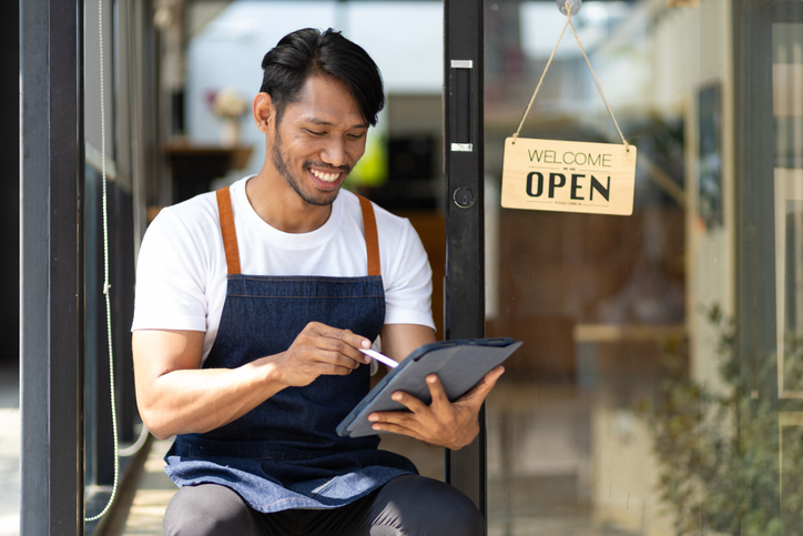 Smiling small business owner wearing an apron sits outside their shop holding a tablet, with an "Open" sign hanging in the background.