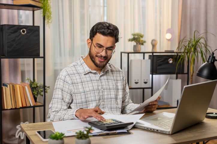 A man with glasses and a plaid shirt works on finances at a desk, using a calculator and laptop while holding a document.